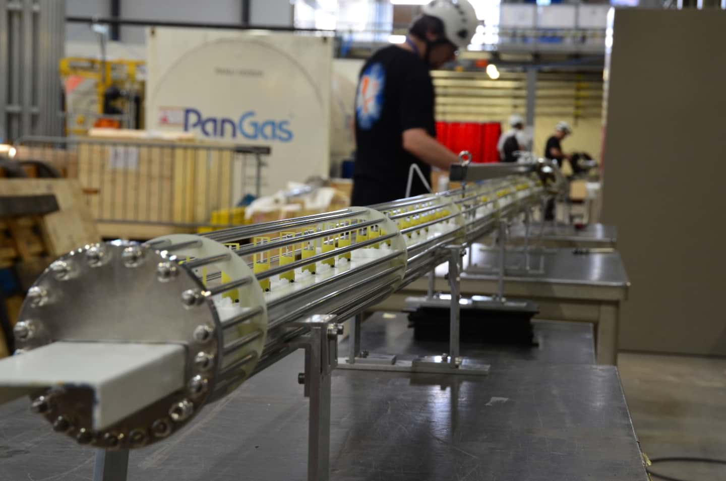 A metallic tube is seen stretching diagonally across the screen. A blurred man in a white hardhat is seen closest to the camera. More people are working further back. A white panel onthe left side has the words PanGas on it.