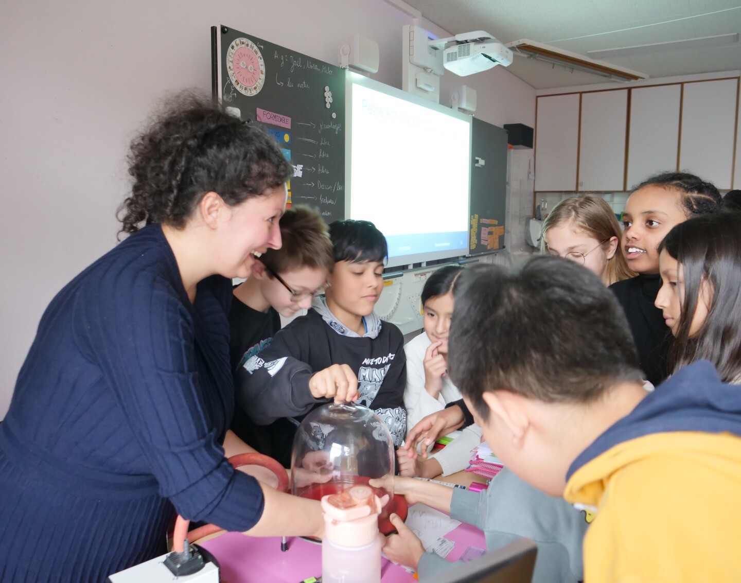 A women engineer surrounded by kids runs a small experiment in a classroom.