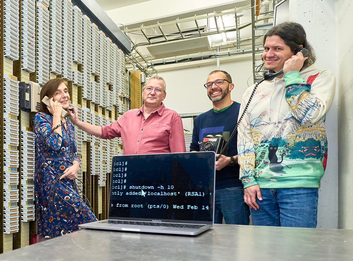 Four people standing in a computer centre, two are holding phones to their ears