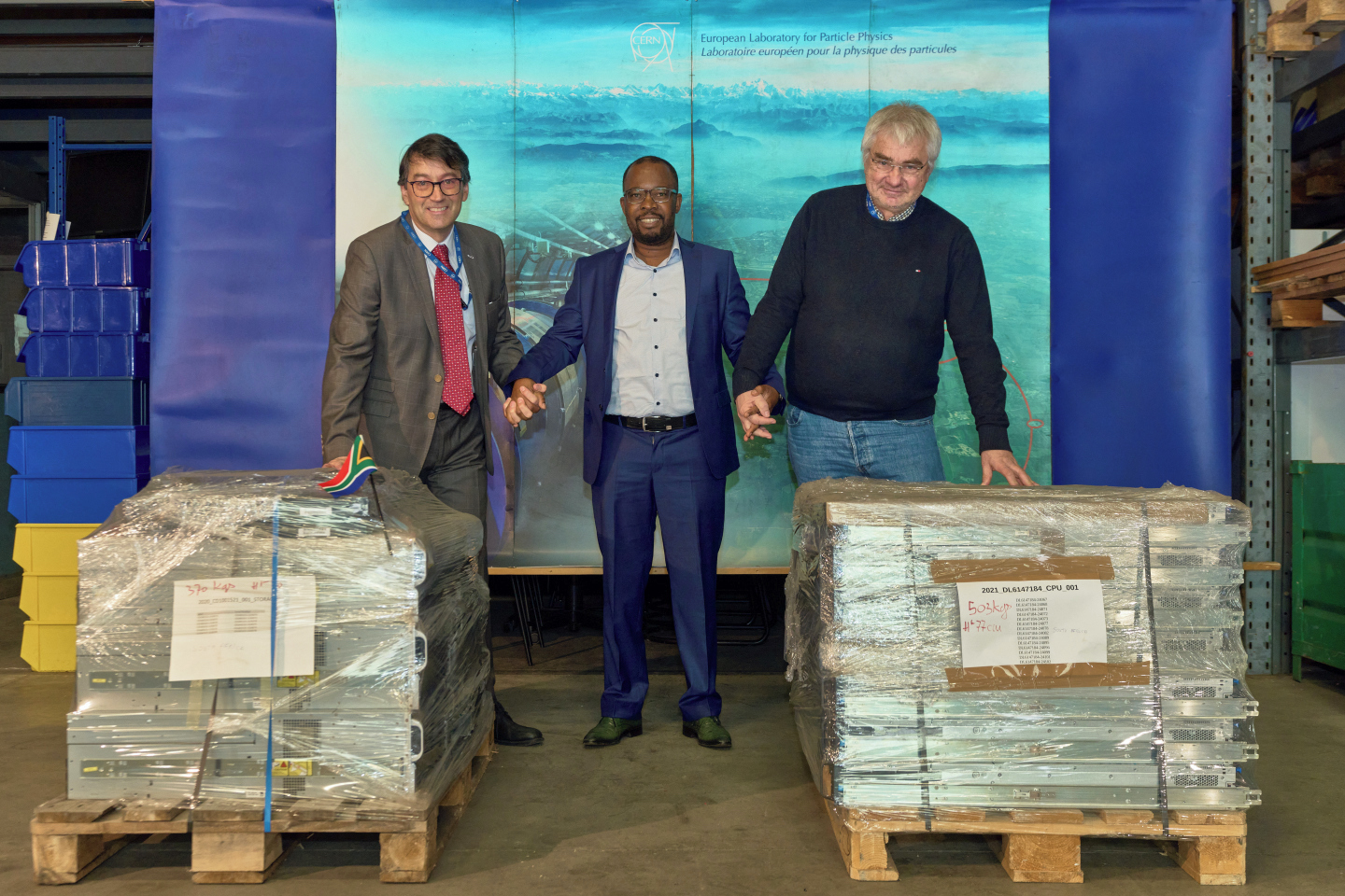 Three men hold hands next to computer hardware packed for shipment