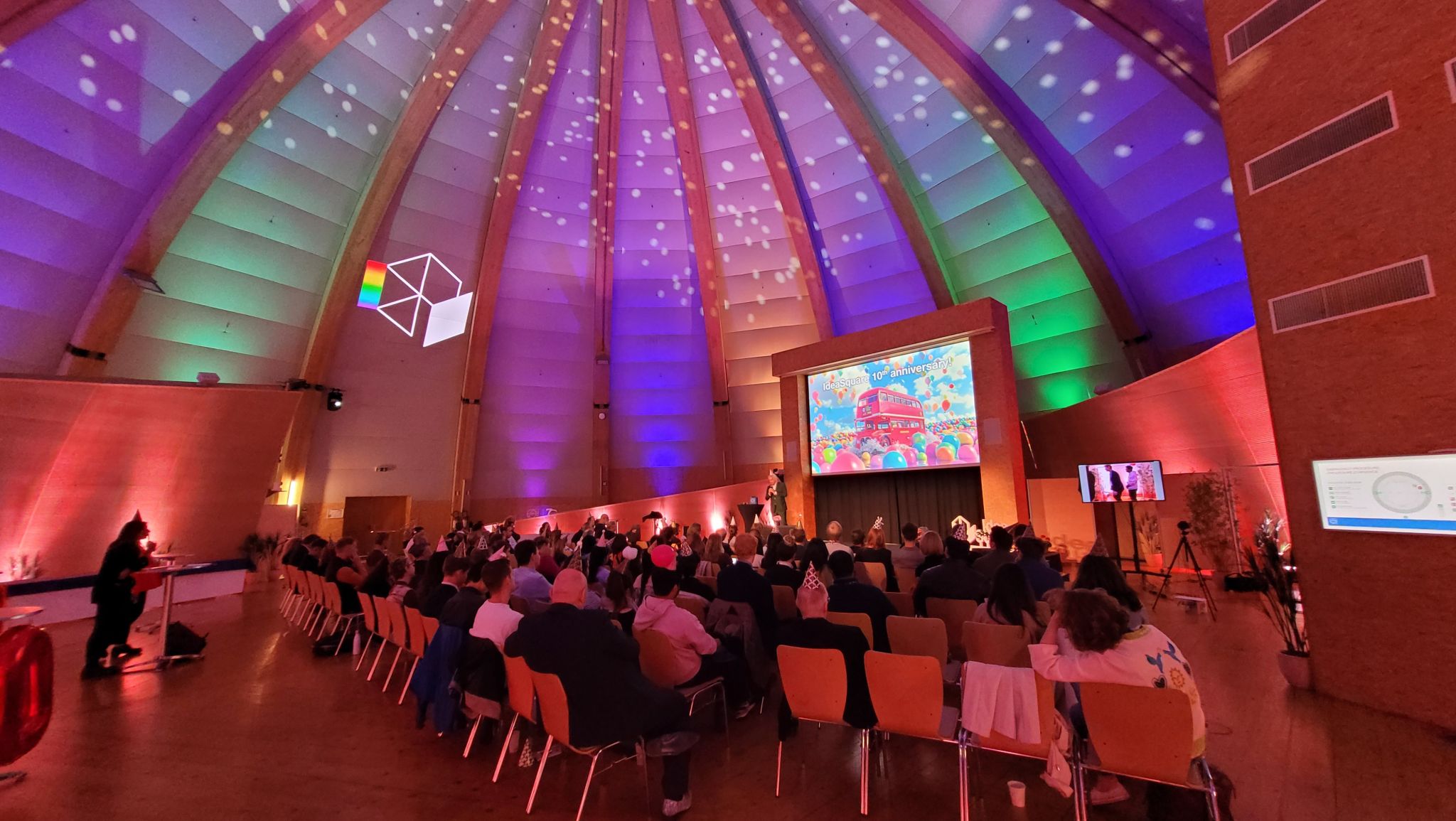 A group of people sat facing a stage, which depicts the IdeaSquare logo and the 10th anniversary poster