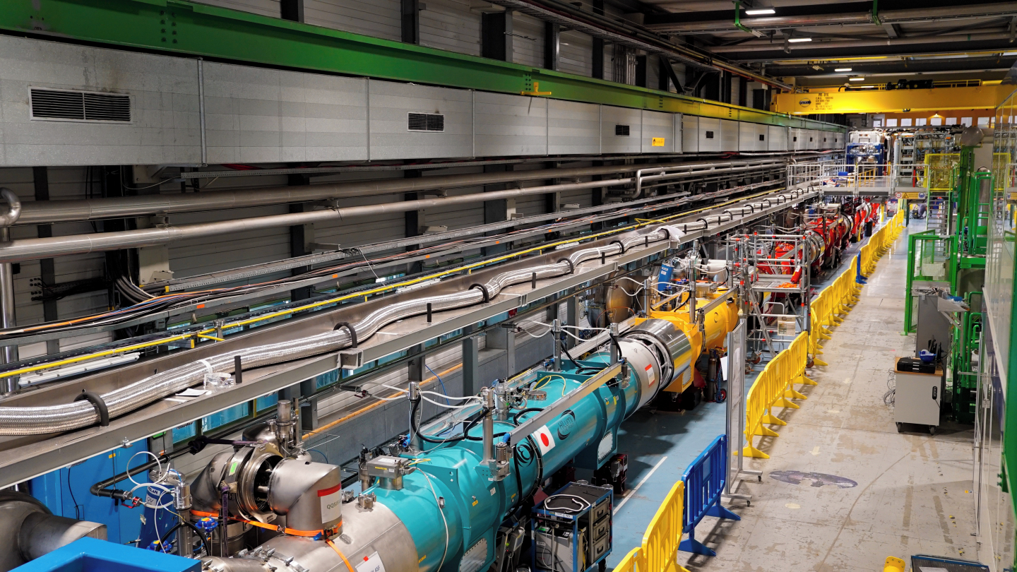 Line of big cylindrical cryostats which house the new magnets. The croystats are blue, rend and yeloow. This line is surrounded by scaffolding and safety barriers.