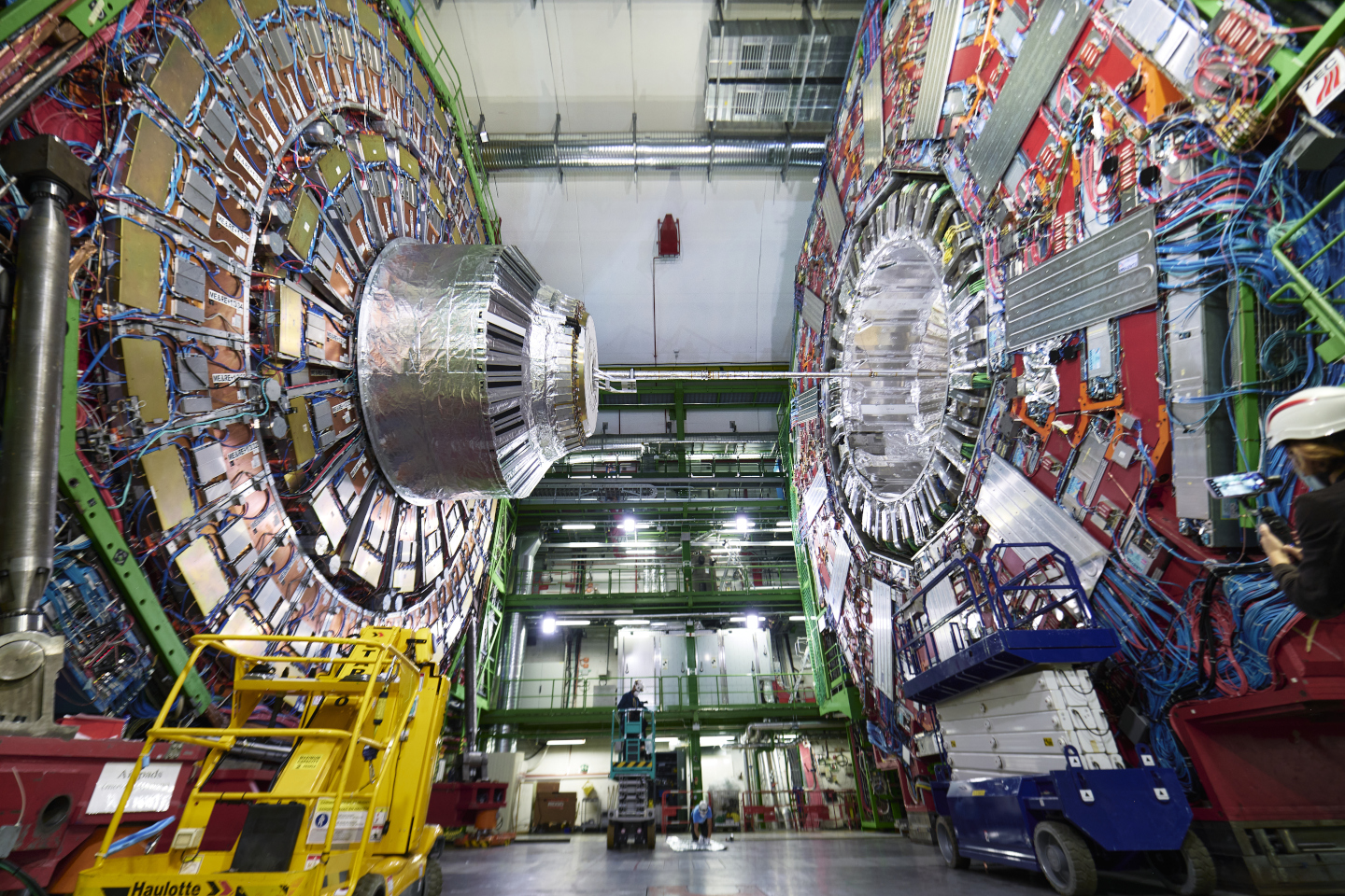 The CMS detector is opened up in a very large hall. The photo is taken from the side and the detector cross-section can be seen on either side of the frame. The detector is metallic and made up of a large number of different brightly-coloured components and wires.