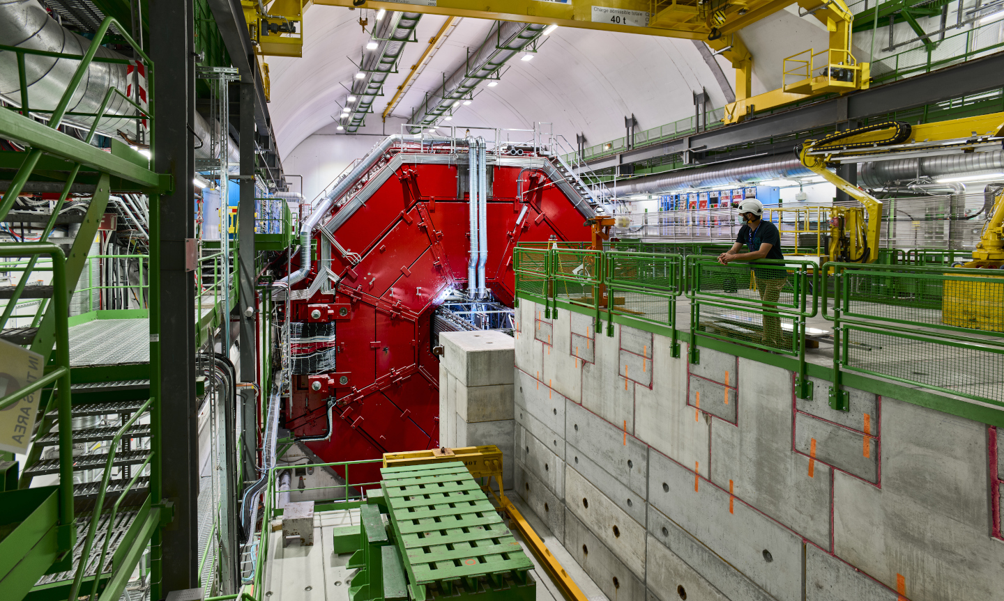 A large cavern in the background is the ALICE detector with the top half of bright red octagonal doors open. IN the foreground is a platform with a person standing on it to give a sense of scale.