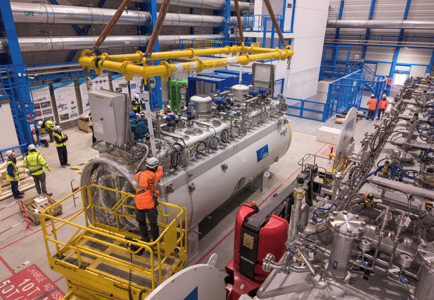 A large hall with concrete walls is filled with large machines with chrome pipes and scaffolding around them. There are people in hi-vis jackets around a new large piece of equipment, the cold box, suggesting it is being installed.