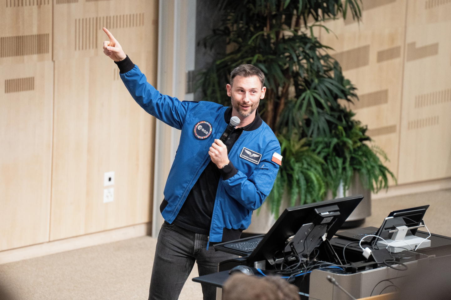 A man speaking into a microphone and pointing up to a screen that is out of view. He is wearing a dark blue jacket with ESA logos.