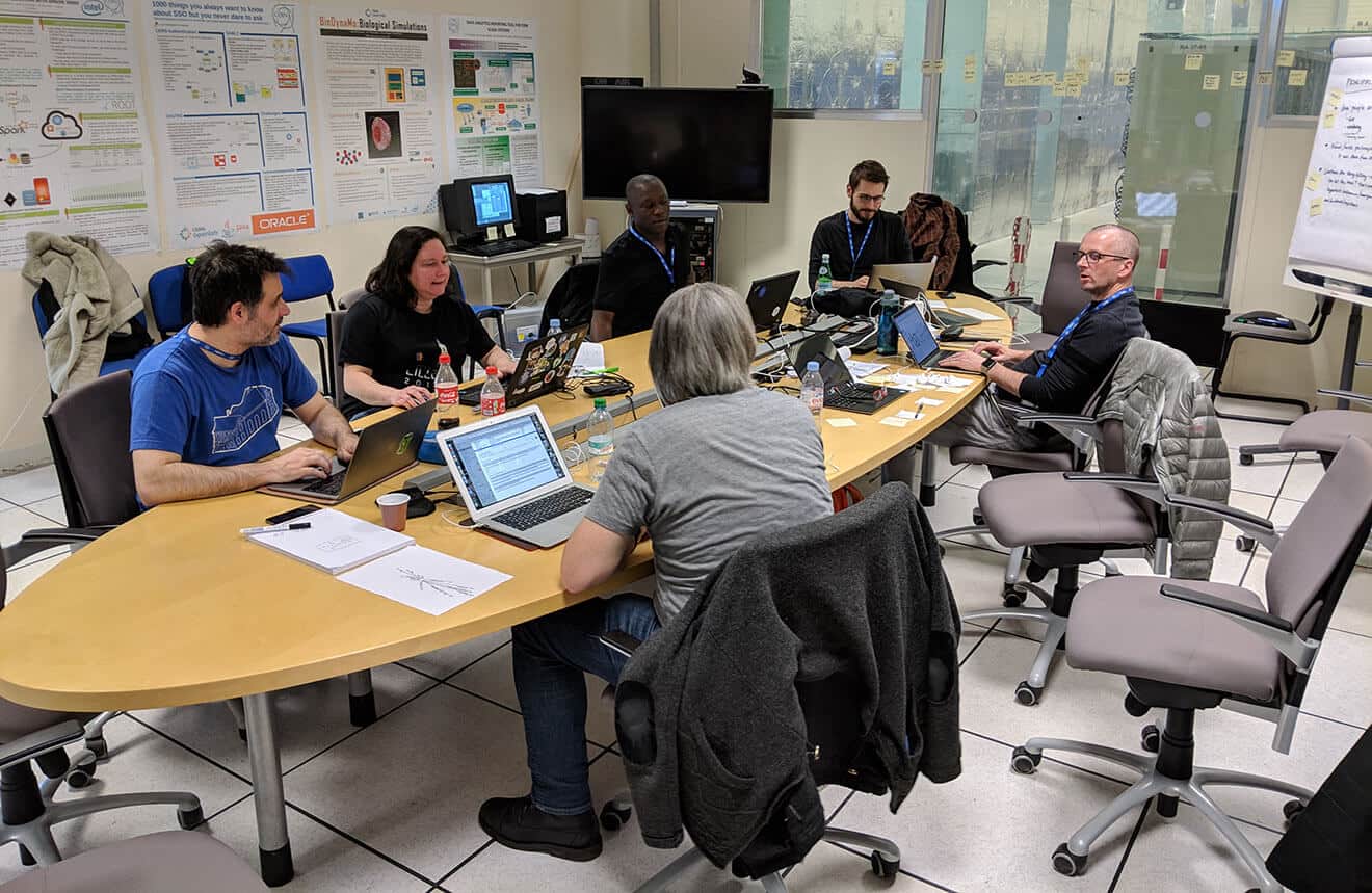 Five men and a woman sit around an oval wooden table with their laptops out. There is a blank screen in the background, and some scientific postersd on the wall. Centred in the image in the background is the NeXT Computer on which the original browser ran